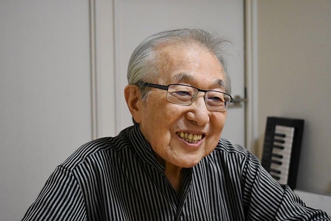 O compositor Chumei Watanabe sorrindo, sentado ao lado de um teclado