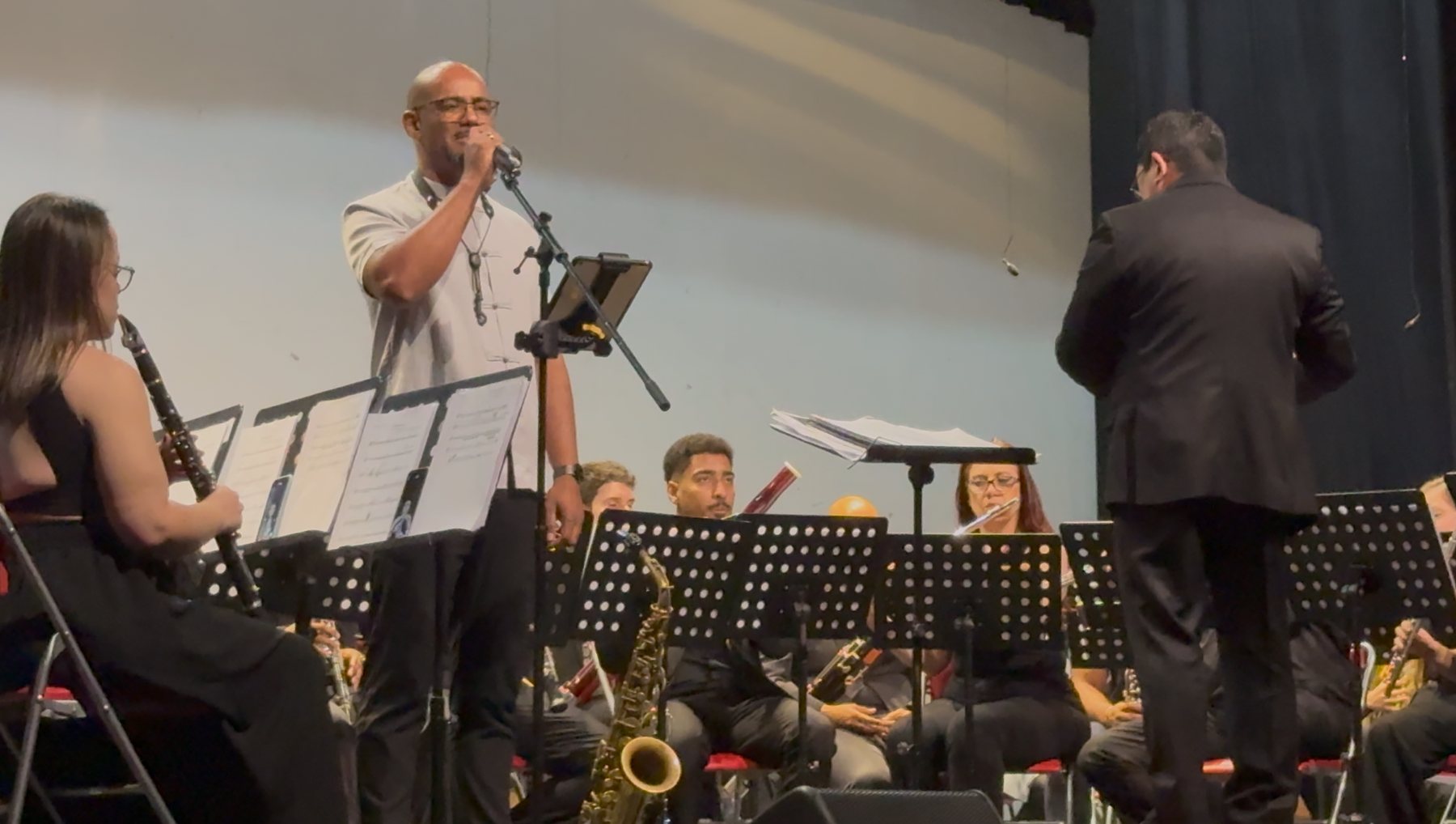 O solista Marcos Andres canta ao microfone diante da Banda Filarmônica de São Paulo, com o maestro de costas à direita