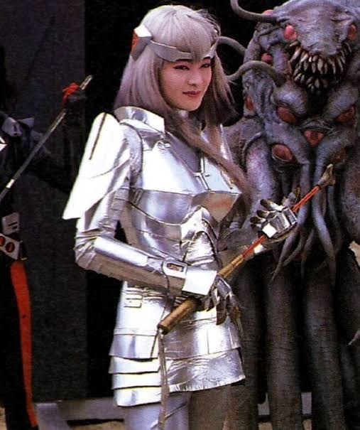 Fukumi Kuroda sorri vestida de Ahames, com a armadura prateada, ao lado de um monstro de várias cabeças com dentes à mostra