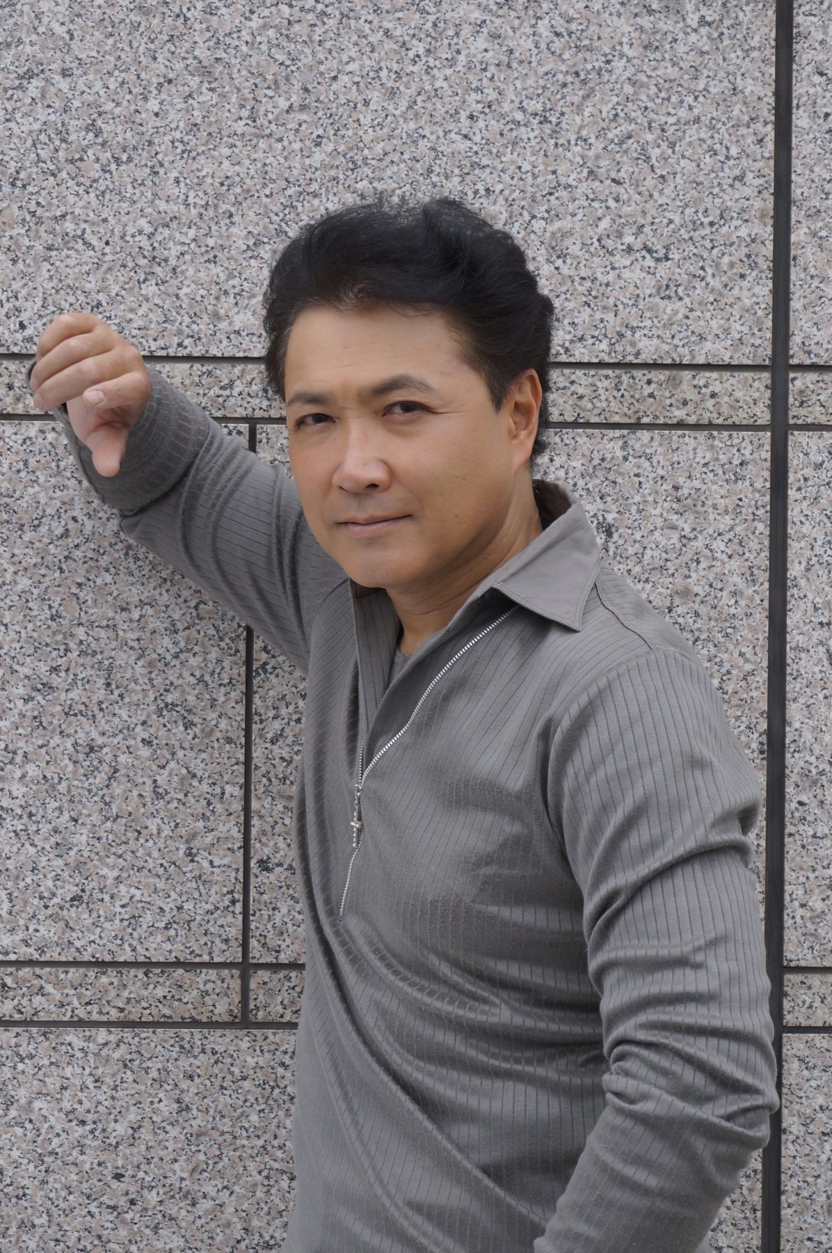 Hiroshi Watari, de blusa cinza com zíper, apoia o braço numa parede de granito