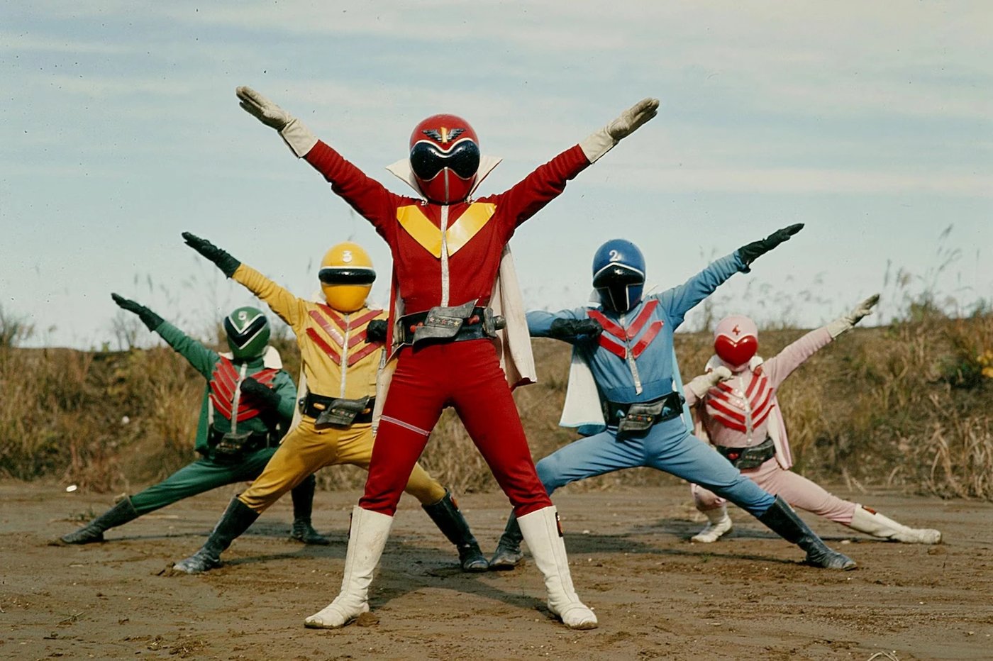 Os cinco heróis de Himitsu Sentai Gorenger fazem a pose de apresentação num terreno de terra, com Aka Ranger ao centro
