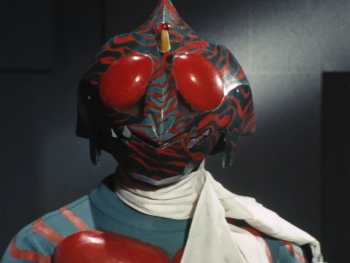 Close da máscara do Kamen Rider Amazon, verde com listras vermelhas e grandes olhos vermelhos, sobre a echarpe branca
