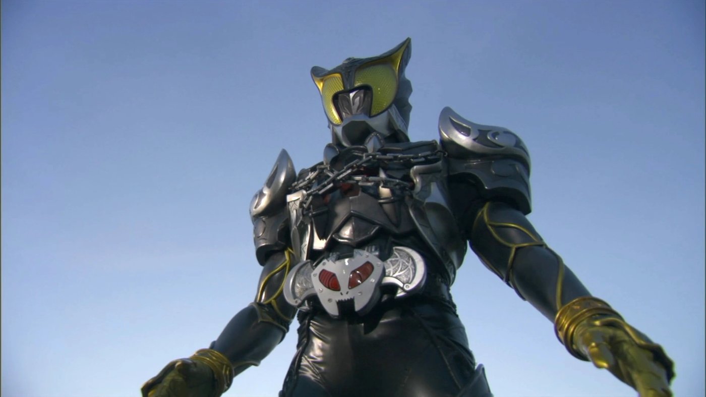 O Kamen Rider Arc visto de baixo para cima contra o céu azul, com correntes cruzadas no peito e viseira amarela