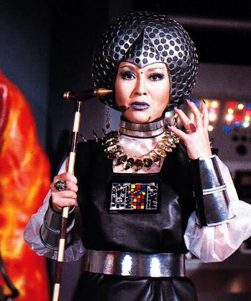 Machiko Soga como a rainha Hedrian de Sun Vulcan, com cetro, capacete prateado e colar de dentes, na base dos vilões