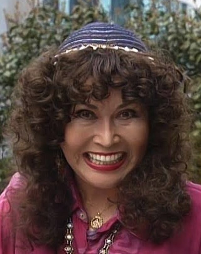 Machiko Soga em cena, de cabelo cacheado, boina roxa e blusa rosa, com um sorriso escancarado