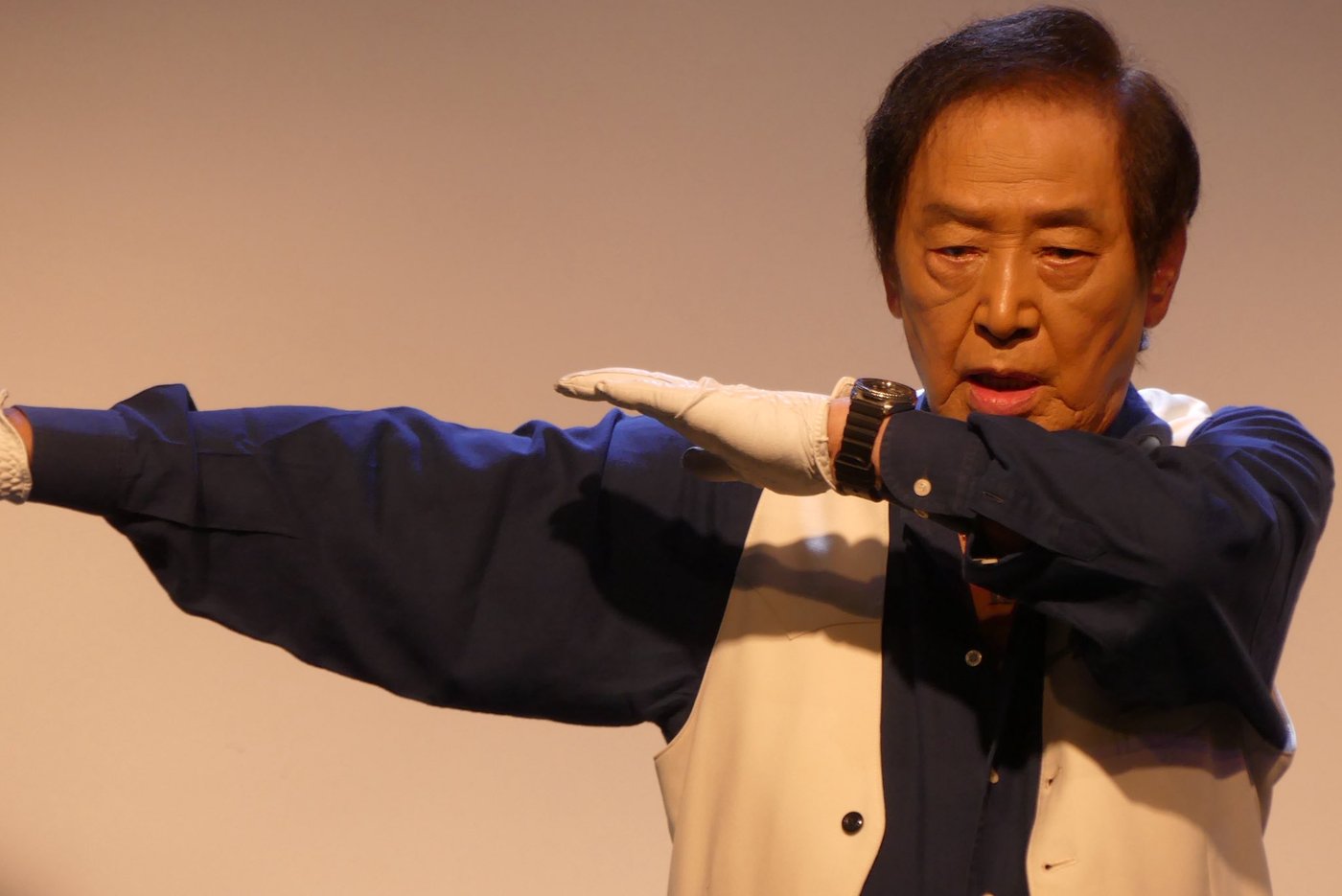 Hiroshi Miyauchi, de luvas brancas e colete branco sobre camisa azul, faz com os braços a pose de transformação de V3