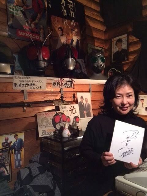 Moyu Taguchi holds a signed shikishi board at Tetsuo Kurata’s bar, decorated with Kamen Rider Black masks and memorabilia