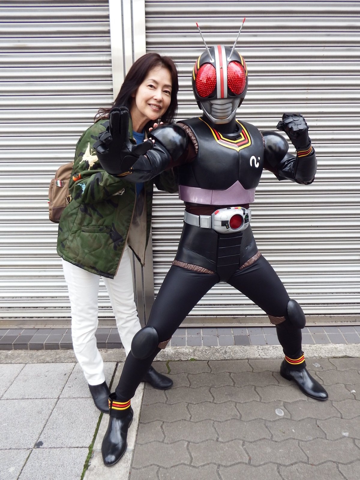 Moyu Taguchi smiles and poses next to a Kamen Rider Black cosplayer in front of a steel shutter