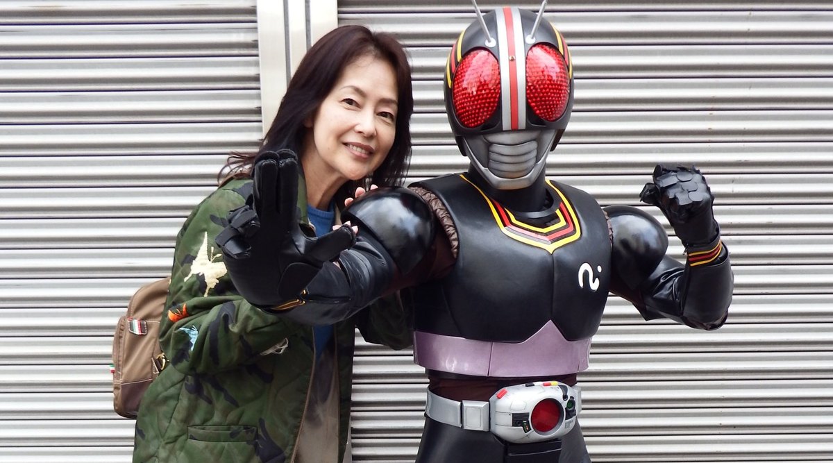 Moyu Taguchi smiles and poses next to Kamen Rider Black in front of a steel shutter