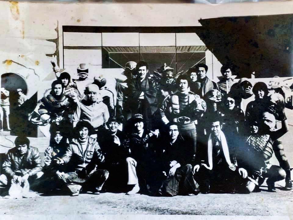 Old black-and-white photo of a large group, with members in hero suits and masks and others in everyday clothes