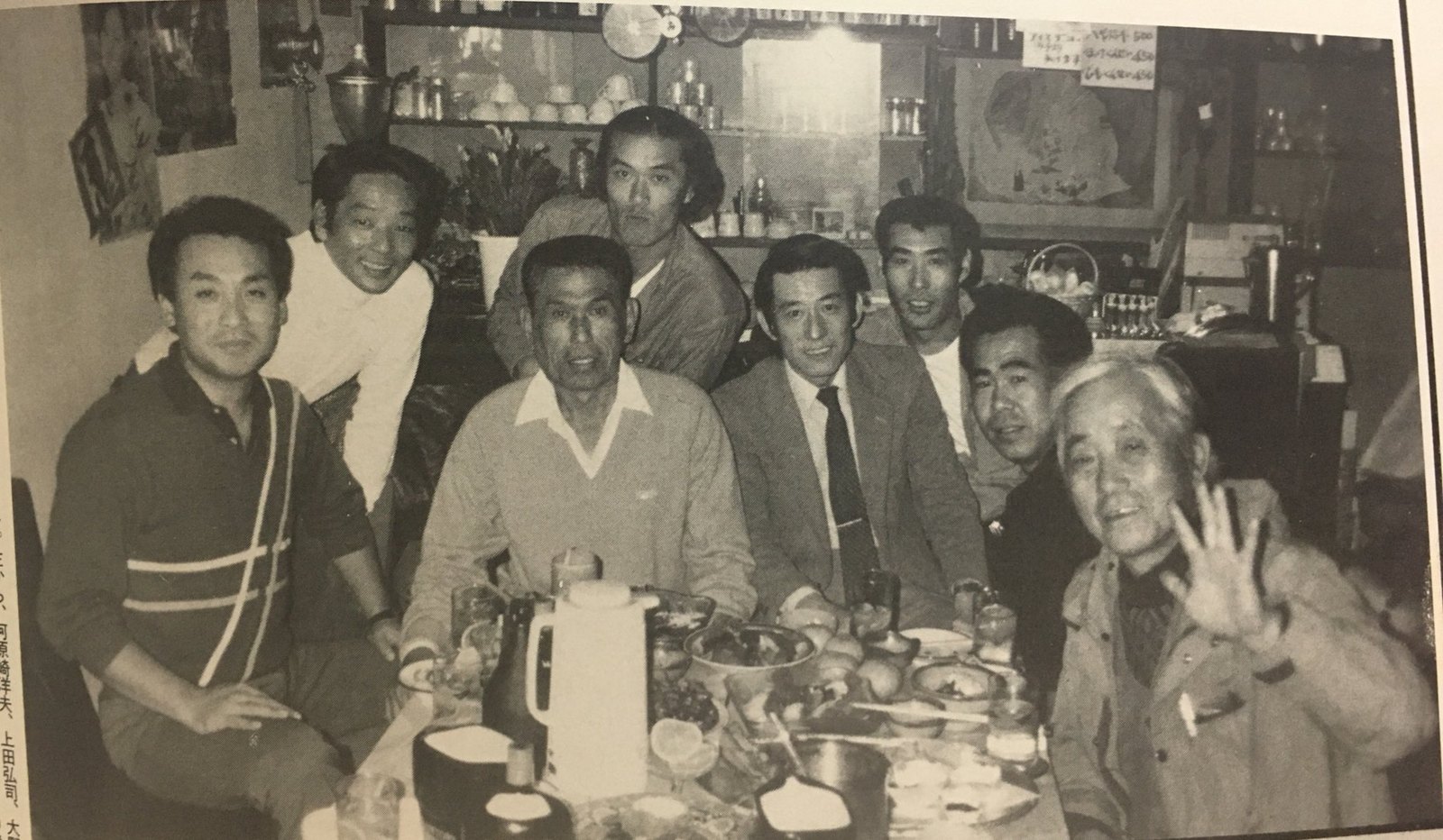 Black-and-white photo of eight team members gathered around a restaurant table, one of them waving