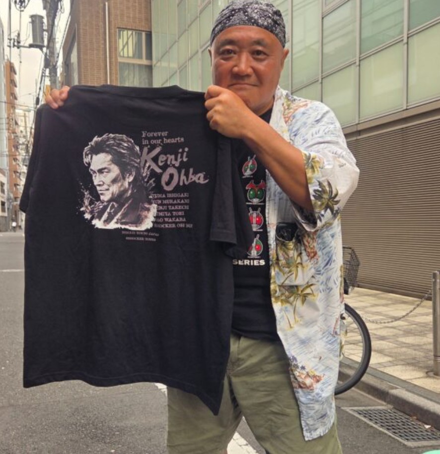 Shocker Ohno holding up a black T-shirt with Kenji Ohba's face and the words Forever in our hearts