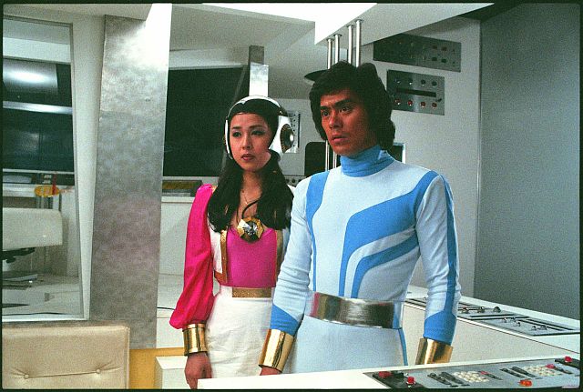 Kenji Ohba, como Retsu Ichijoji, de uniforme branco e azul, ao lado de Wakiko Kano, como Mimi, de rosa, numa sala de comando
