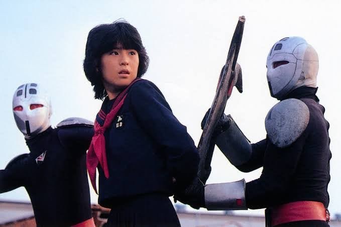 Tokie Shibata, de uniforme escolar de marinheiro e lenço vermelho, olha para trás entre dois soldados de máscara prateada e olhos vermelhos