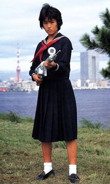 Tokie Shibata como Nana, de uniforme escolar de marinheiro e lenço vermelho, segura a arma de Rigel à beira de uma baía, com a Torre de Tóquio ao fundo