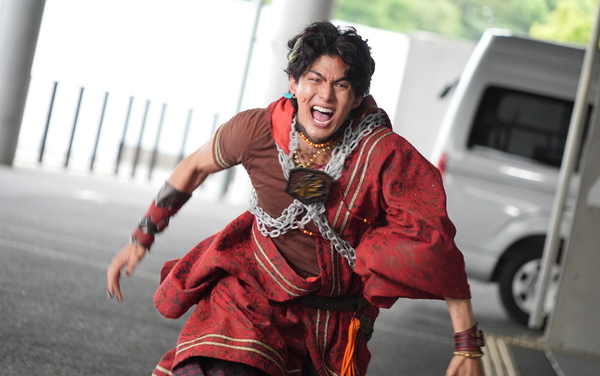 Personagem de túnica vermelha, com correntes cruzadas no peito, corre em direção à câmera em cena de Kakusei Hunter Omegahorn