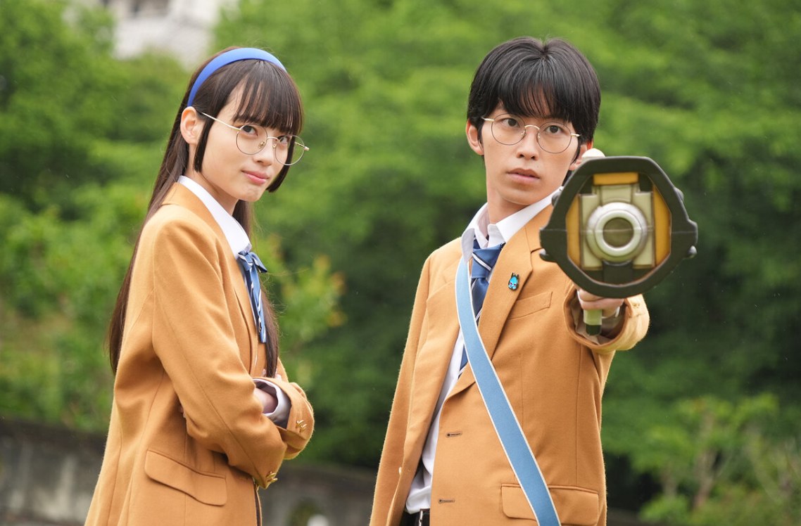 Dois personagens de uniforme escolar e óculos em cena de Kakusei Hunter Omegahorn; um deles aponta um aparelho para a frente