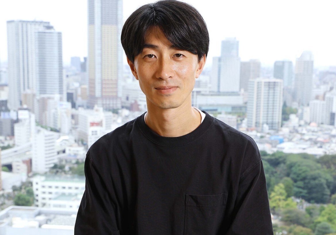 O produtor Yoshito Kuji, de camiseta preta, diante de uma janela com vista para os prédios de Tóquio