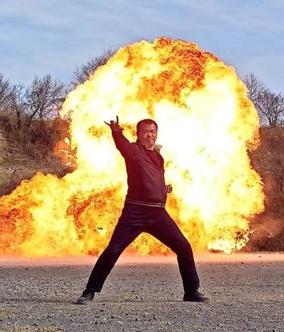 素顔の岡元次郎が爆発を背にヒーローのポーズを取る
