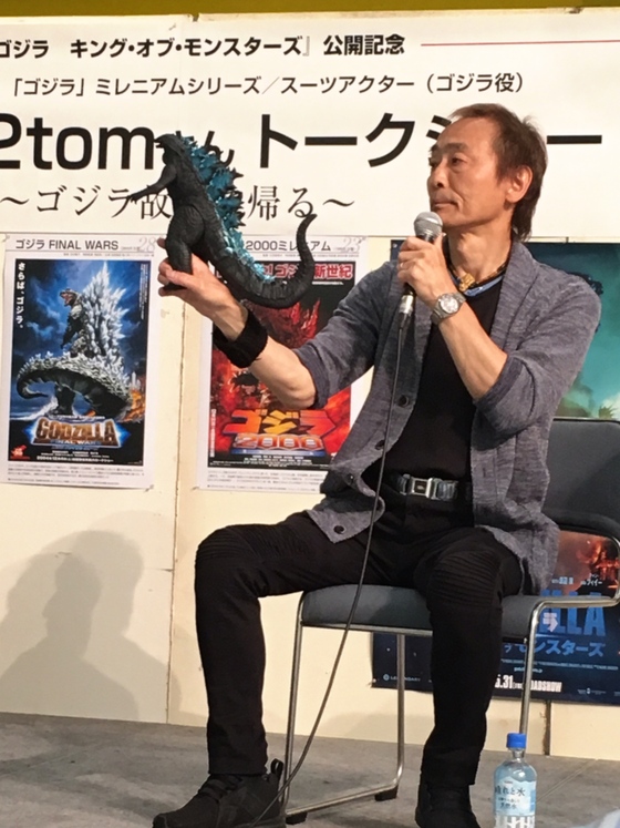 Kitagawa segura uma figura de Godzilla em talk show, entre pôsteres da era Millennium